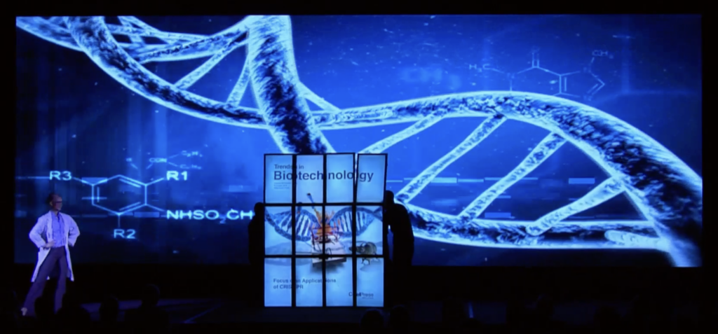 A person stands on stage with a large screen displaying a DNA double helix and chemical formulas in the background. The words Biotechnology and Frontiers & Applications of CRISPR are on a large book or screen in the center.